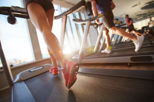 Low-angle View Of Male And Female Legs Running On Treadmill In Gym On Sunny Day