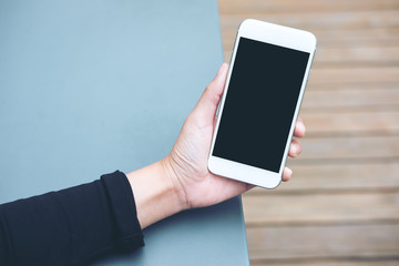 Mockup image of hands holding white mobile phone with blank black screen on table and wooden floor background 