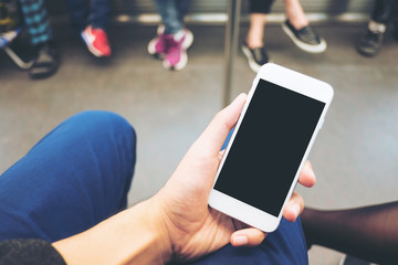 Mockup image of hand holding white mobile phone with blank black screen in subway with many people in background
