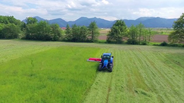 A man is sitting in a blue tractor and he is working out in the fields. He is cutting grass with the help of a big agricultural machinery.
