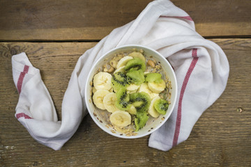 From above view of white ceramic bowl of muesli with soy milk and slices of fresh banana and kiwi fruits on wooden table with white towel