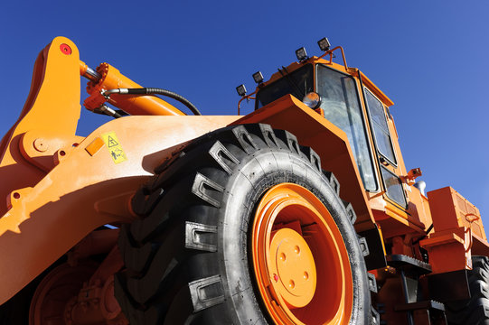 Bulldozer, huge orange powerful construction machine with hydraulic piston of scoop and black wheels, heavy industry, bottom view, blue sky on background 