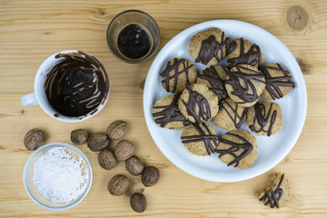 Freshly baked homemade walnut cookies with chocolate icing on a plate surrounded by baking ingredients and whole nuts, overhead view