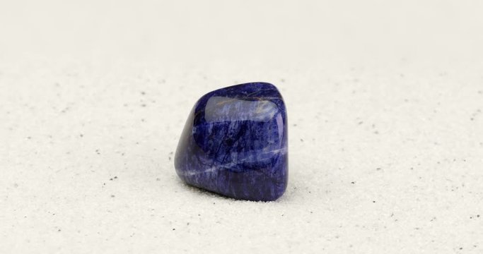 Sodalite gem on white sand surface. Tumbled semi precious gemstone rotating in seamless loop.
