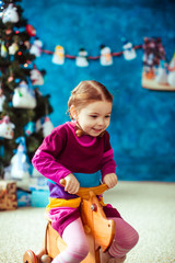 Smiling little girl sits on wooden horse before Christmas tree