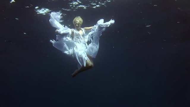 Underwater model free diver in costume angel swims in clean water in Red Sea. Filming a movie. Young girl smiling at camera. Extreme sport in marine landscape, coral reefs, ocean inhabitants.