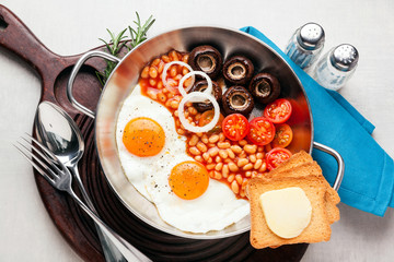 Breakfast with fried eggs in cast-iron pan, broken egg, tomatoes, on cutting board over grey stone background. Top view with copy space