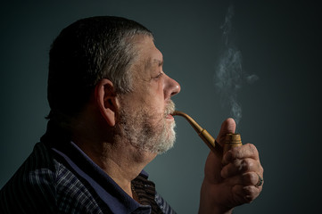 Film toned portrait of Caucasian bearded man smoking tobacco pipe against dark background.