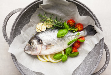 Fresh uncooked dorado or sea bream fish with lemon, herbs, oil, vegetables and spices in a frying pan on a baking paper over white backdrop, top view