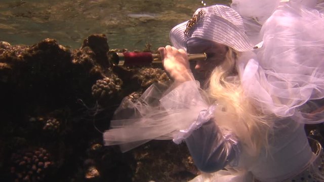 Model in white costume of pirate underwater in Red Sea. Filming a movie. Young girl smiling at camera. Extreme sport in marine landscape, coral reefs, ocean inhabitants.