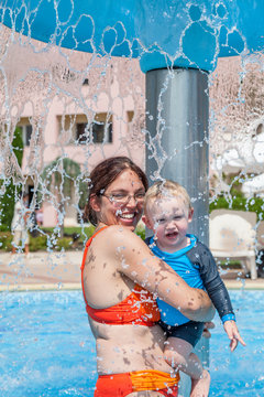 Mother and child together in water fall shower in pool outdoors. Summer vacation resort.
