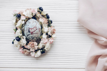 happy easter greeting card. easter egg with chick ornaments in floral nest with flowers on rustic white wooden desk background. space for text, top view. soft light