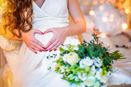 Pregnant girl in white dress, her hands on her stomach in form of heart
