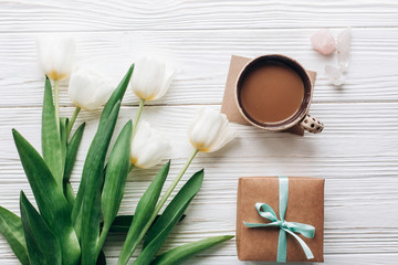 stylish craft present with morning coffee and tulips on white wooden rustic background. boho flat lay with flowers and gift with space for text. hello spring concept