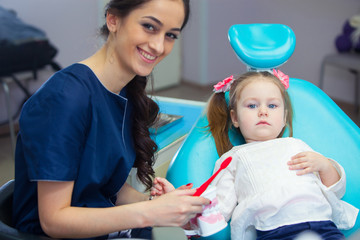 Pediatric dentist educating a smiling little girl about proper tooth-brushing, demonstrating on a model. Early prevention, raising awareness, oral hygiene demonstration concept.