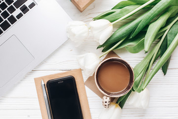 phone screen and laptop with morning coffee and tulips on white wooden rustic background. stylish flat lay with flowers and working gadgets with space for text. freelance. home working