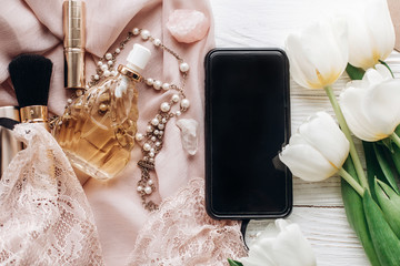 Obraz na płótnie Canvas phone screen and stylish woman lace lingerie jewelry and perfume present on soft fabric and tulips on white rustic background. flat lay girl essentials for a holiday. sensual photo