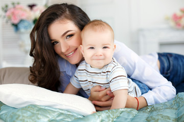 Happy mother, woman, brunette with long curly hair, has fun and plays with his young son,the kid with blond short hair,wearing a white striped T-shirt and blue jeans,lying on a bed of turquoise color