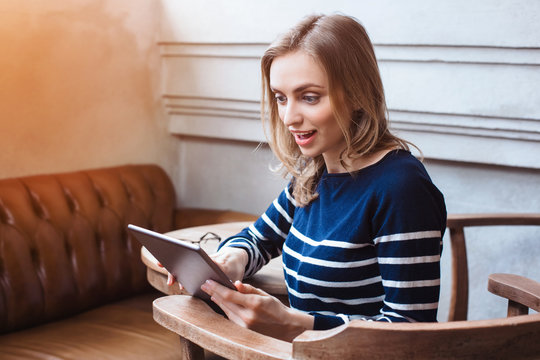 Young smaling female student is chatting on tablet pad with friend while sitting in modern cafe, gorgeous Caucasian woman using digital tablet while rest in cafe during spring holidays - Powered by Adobe