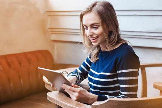 Young smaling female student is chatting on tablet pad with friend while sitting in modern cafe, gorgeous Caucasian woman using digital tablet while rest in cafe during spring holidays