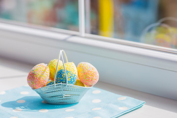 Colorful easter eggs in white basket on blue fabric near window