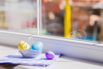 Colorful easter eggs in white basket on blue fabric near window - 139012697
