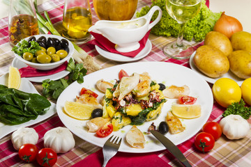 Grilled calamari on a white plate with boiled potatoes and chard, lemon, cherry tomatoes and olives, served next to fresh vegetables and sauce.