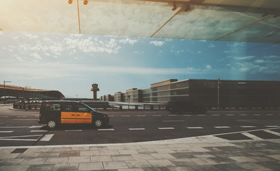 Yellow taxi on the road in front of air traffic control tower of modern contemporary airport terminal of Barcelona with buses and multiple moving blurred cars in distance on sunny day