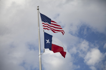 Naklejka premium American and Texas Flags Waving Together Against Blue Sky with Clouds