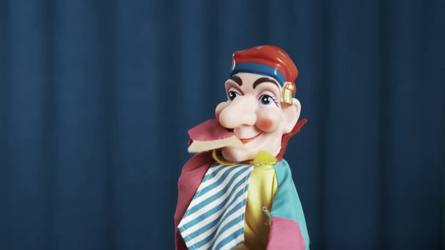 Funny jester hand puppet toy shake hand and put it to mouth, nodding on scene with blue crease curtains background