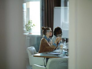 Side view of modern young people in casual clothes sitting at meeting table in conference room...