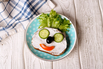Kids meal, breakfast or lunch for children. Colorful funny food face with healthy vegetables, cheese spread and green salad on a plate. Concept of healthy food, healthy breakfast. Top view, closeup