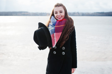 Funny casual young girl at black coat, scarf and hat against frozen river on sunny winter weather.