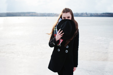 Funny casual young girl at black coat, scarf and hat against frozen river on sunny winter weather.