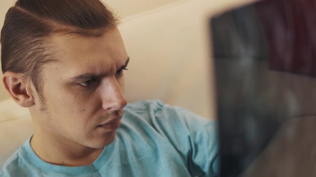 Young caucasian concerned man in blue shirt with hair knot sitting on  white leather  sofa observes printed x-ray sheet, close up