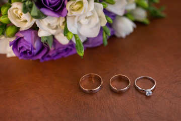 Two golden wedding rings and engagement ring with a diamond on brown background with white and purple flowers