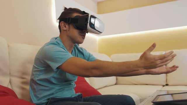 Young caucasian naughty man wears virtual reality headset in blue shirt with hair knot sitting on white leather sofa in modern apartment living room playing lewd video game.