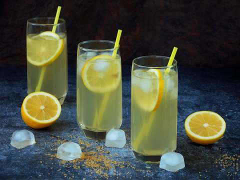 Three glasses of cold homemade lemonade with lemon slices, ice cubes, brown sugar and straws on dark background. Copy space