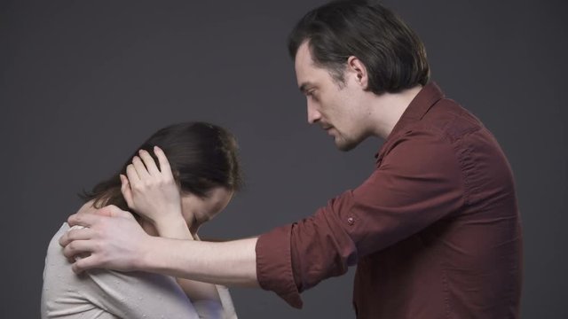 Young couple having a quarrel, woman is crying, a man holding her shoulders and waving her, gray background