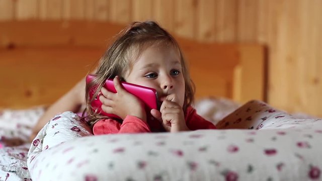 Little girl lies on the pillow and holds in hands mobile phone. Little girl with red cell phone lies on the sofa. Funny little girl with red smartphone lies on the bed and picks his nose