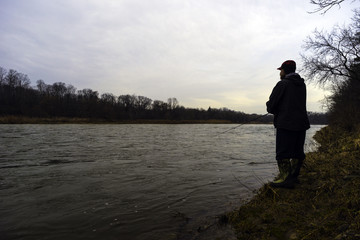 Fototapeta na wymiar Early spring fisherman fishing off bank of fast flowing river at sunrise