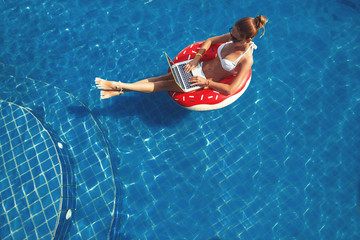 Pretty female in swimsuit and sunglasses sitting in swimming ring in pool, enjoy and using laptop....