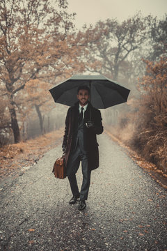 Businessman con giacca guanti e ombrello nero e borsa marrone sorride in una via di campagna durante la pioggia.