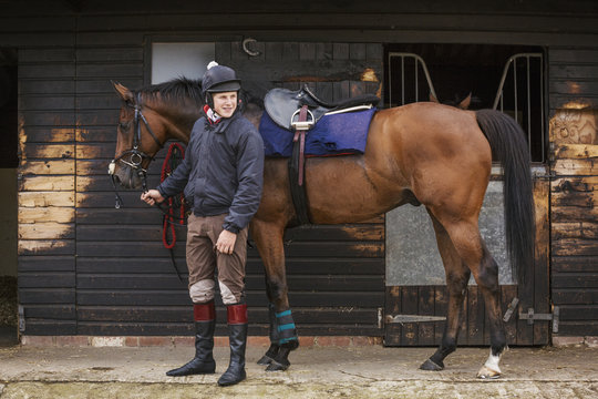 Man wearing riding gear standing outside a box stall at stable with bay horse