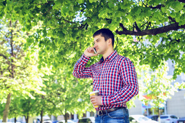 Man talking on the phone, in the summer in the city park, a lot of green trees, smiling happily. Outdoors. The glasses and a shirt with a coffee or tea in hand.