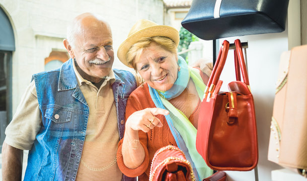 Senior Couple Shopping At Fashion Bag Store With Wife Pointing Showcase To Husband - Active Elderly Concept With Mature Man And Woman Having Fun In City - Happy Retired People Moments On Vivid Colors