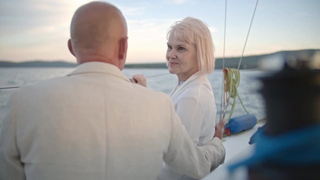 Attractive senior lady sitting on deck of sailboat on yacht and listening to her husband
