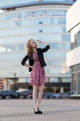 Beautiful blonde with curly hair girl in a dress and black leather jacket stands in the background of the business center with reflective glass windows reflecting the sun. In full growth.