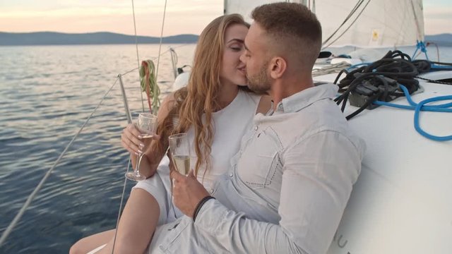 Young couple sitting on edge of yacht, holding glasses of wine and kissing