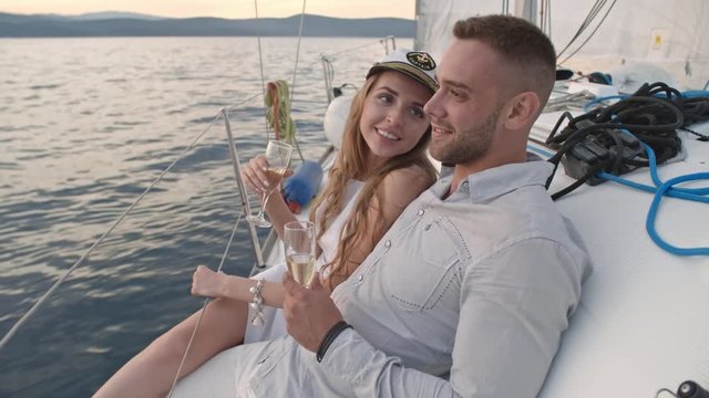 Beautiful young woman in sailor cap sitting aboard with her boyfriend. They doing champagne toast and sipping from flutes while having yacht cruise